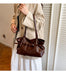 A person wearing a beige trench coat and white skirt carries a glossy dark brown leather shoulder bag with a structured top handle and a front strap detail, standing indoors against a white slatted door.