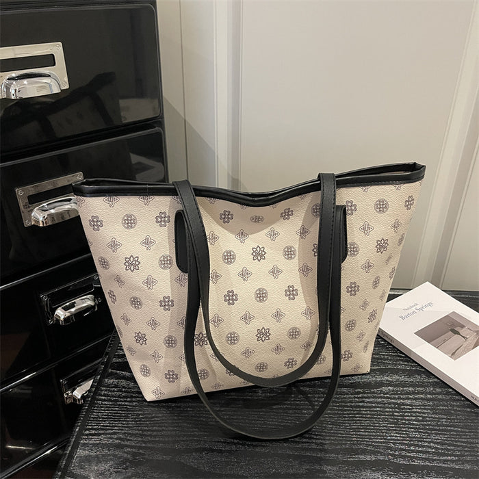 A beige canvas tote bag with a repeating geometric monogram pattern and black leather trim, handles, and straps, displayed on a dark wooden surface next to a black drawer and a book.