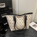 A beige canvas tote bag with a repeating geometric monogram pattern and black leather trim, handles, and straps, displayed on a dark wooden surface next to a black drawer and a book.