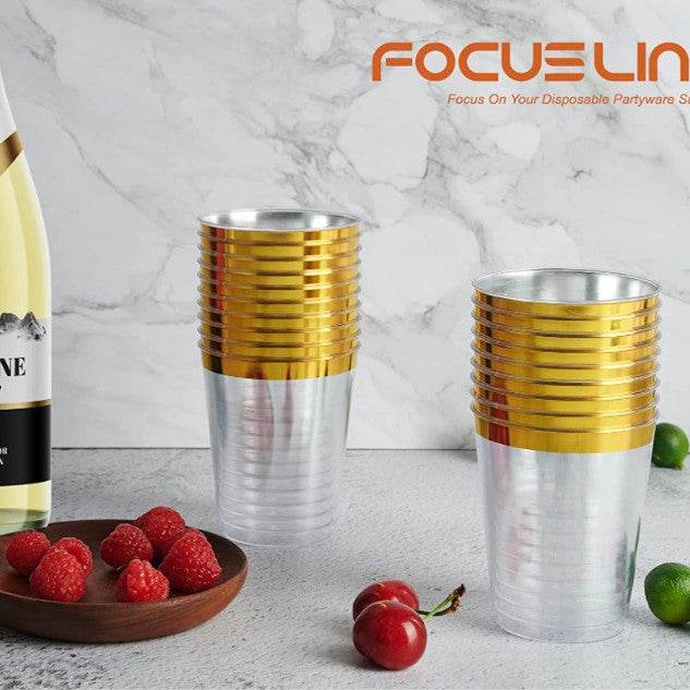 Stacks of gold and silver disposable party cups on a marble surface with a bottle of wine and fruits.