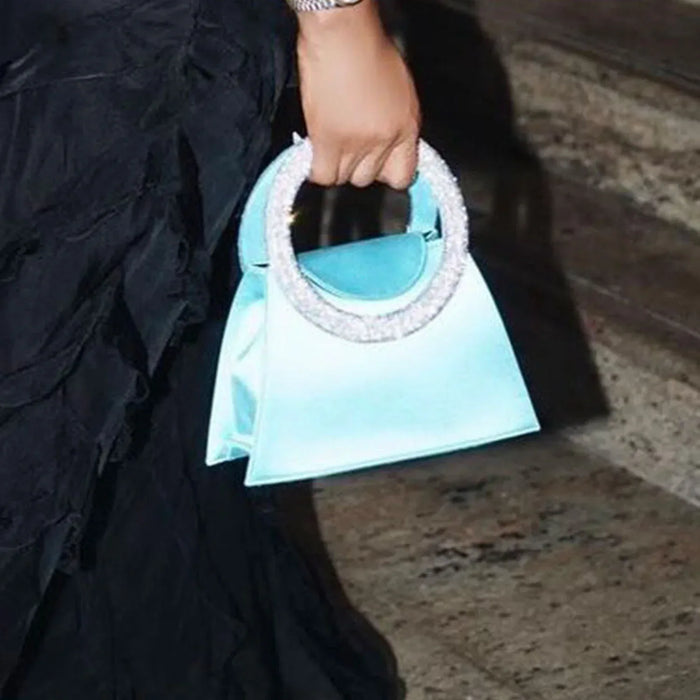 Hand holding a light blue handbag with decorative handle on a dark background
