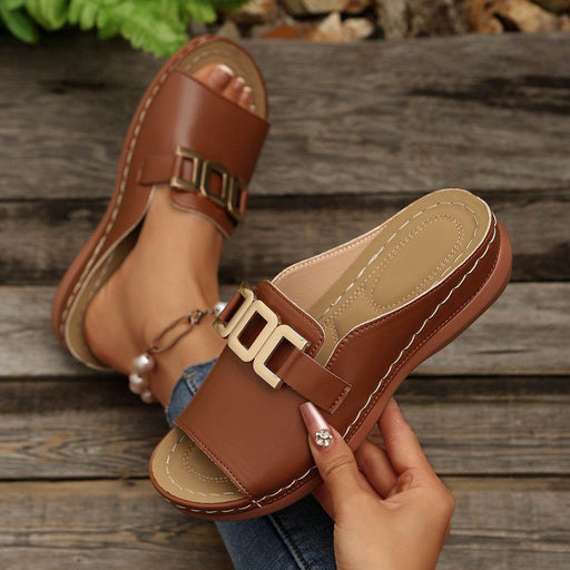 Brown sandals with gold buckles being held by a hand on a wooden surface.