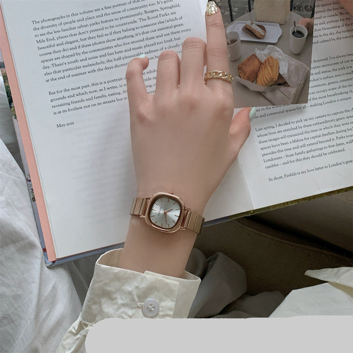A close-up of a person's hand wearing a rose gold-toned watch with a round white dial, resting over an open book with printed text and a small food photo.