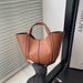 A tan leather tote bag with black piping and double top handles, held by a hand wearing a dark gray sleeve in front of window blinds.