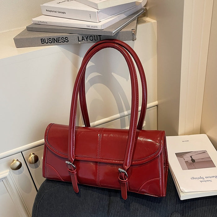 A glossy red leather handbag with double top handles and silver-tone hardware, styled on a dark textured surface next to books in an indoor setting.