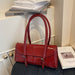 A glossy red leather handbag with double top handles and silver-tone hardware, styled on a dark textured surface next to books in an indoor setting.
