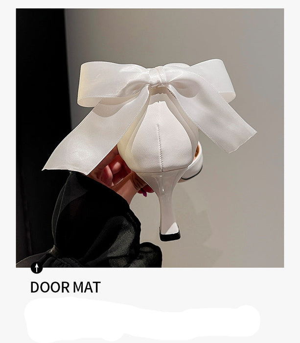 White high-heeled shoes with a large bow held by a hand against a neutral background.