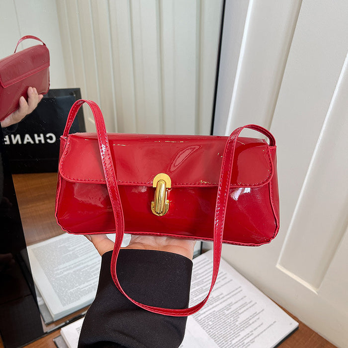 A red patent leather shoulder bag with a gold turn-lock clasp, held in a hand. A Chanel shopping bag is visible in the background on a wooden table.
