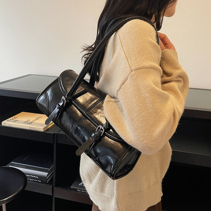 A woman wearing a beige knit sweater carries a shiny black leather shoulder bag with a cylindrical shape and multiple straps, standing indoors near a dark shelf.