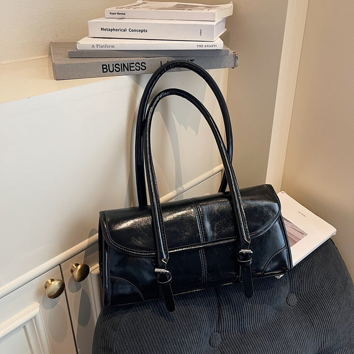 A black leather handbag with double top handles and a flap closure, styled on a dark tufted bench next to a white cabinet with books on a shelf.