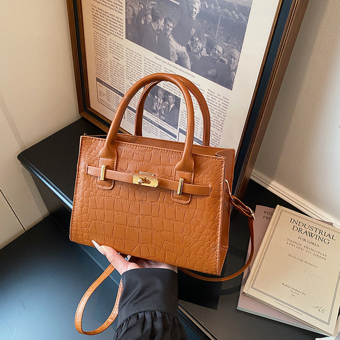 A tan crocodile-embossed leather handbag with double top handles and a gold-tone front clasp, held by a hand in a black sleeve. The bag is styled in an indoor setting next to a framed print and a book.