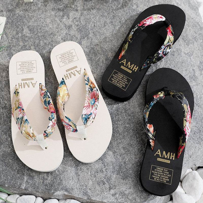 Pair of white and black floral flip-flops with 'AMH' branding on a stone surface.