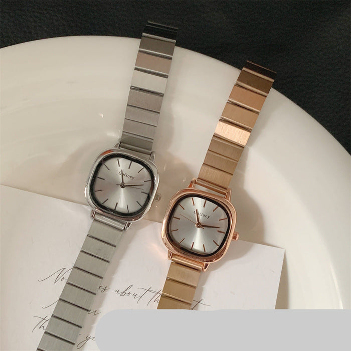 Two analog wristwatches with rounded square faces and metal link bands, one in silver and one in rose gold, displayed on a white plate with a card underneath.