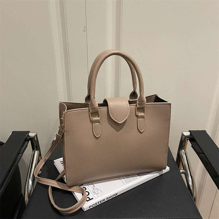 A structured taupe leather tote bag with double top handles and a shoulder strap, resting on a black table with a magazine.