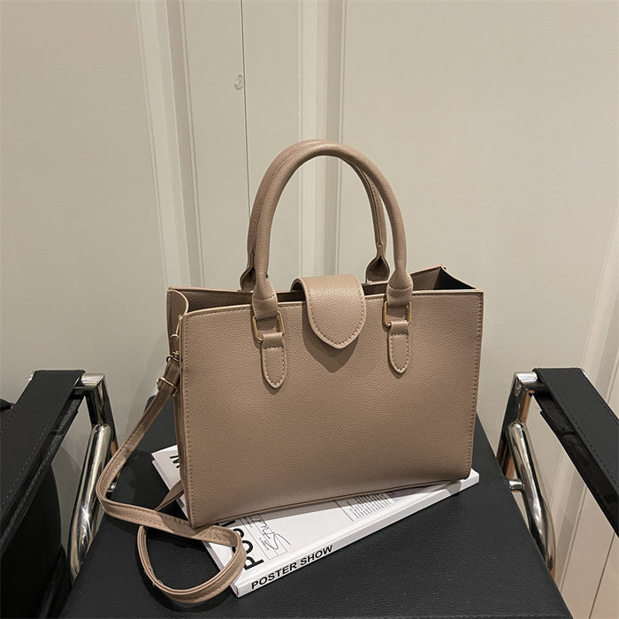 A structured taupe leather tote bag with double top handles and a shoulder strap, resting on a black table with a magazine.