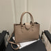 A structured taupe leather tote bag with double top handles and a shoulder strap, resting on a black table with a magazine.