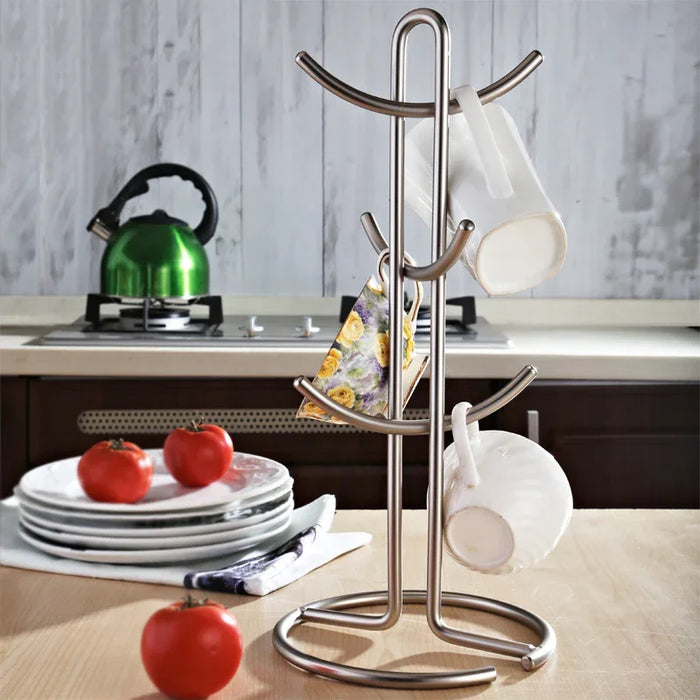 Stainless steel mug tree on a kitchen counter with tomatoes and a kettle in the background.