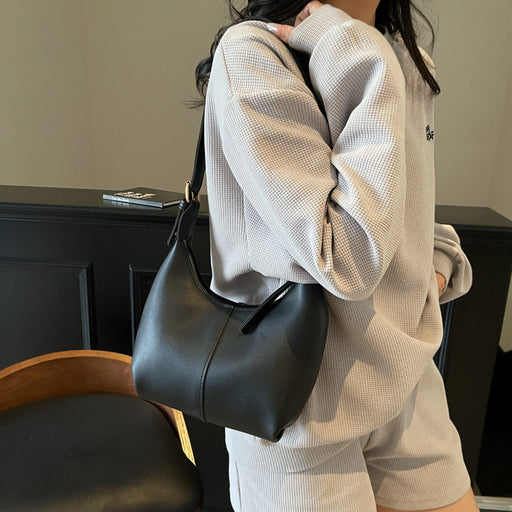 A person wearing a light beige waffle-knit sweatshirt and shorts carries a black leather shoulder bag with a slouchy shape and short strap.