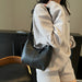 A person wearing a light beige waffle-knit sweatshirt and shorts carries a black leather shoulder bag with a slouchy shape and short strap.