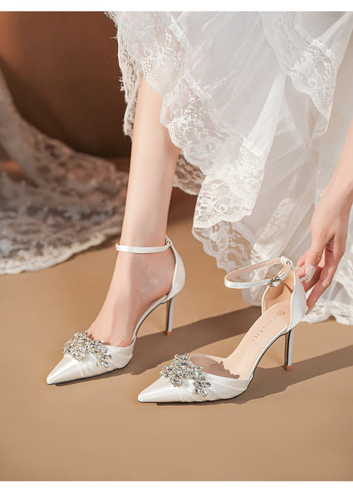 White high-heeled shoes with embellishments worn by a person in a white lace dress.