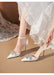 White high-heeled shoes with embellishments worn by a person in a white lace dress.