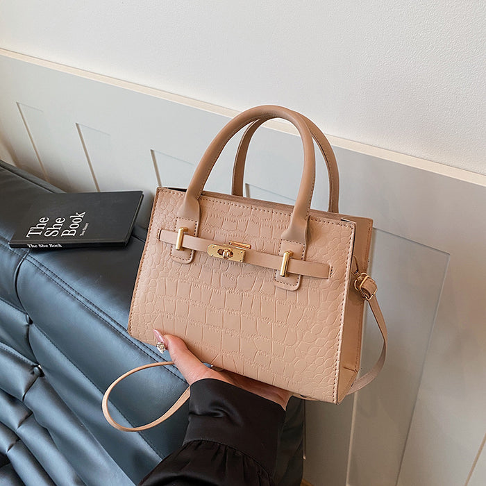 A structured beige handbag with a crocodile-embossed texture, dual top handles, and gold-tone hardware, held by a person wearing a black sleeve in front of a gray wall.