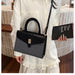 A woman in a white ruffled blouse carries a structured handbag with a black top handle and flap, featuring a textured dark gray lower body and a gold-tone front clasp. She is holding a black book in a tiled indoor setting.