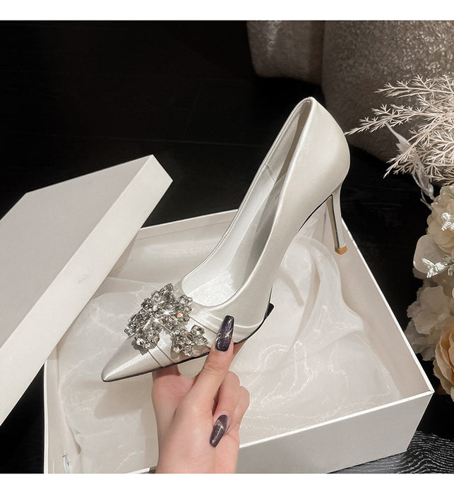 Silver high-heeled shoe with a decorative brooch held by a hand over a white box.