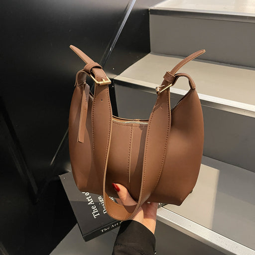 A brown faux leather slouchy shoulder bag with a top zipper and adjustable shoulder straps, held by a hand with red nail polish against a modern black and gray staircase background.