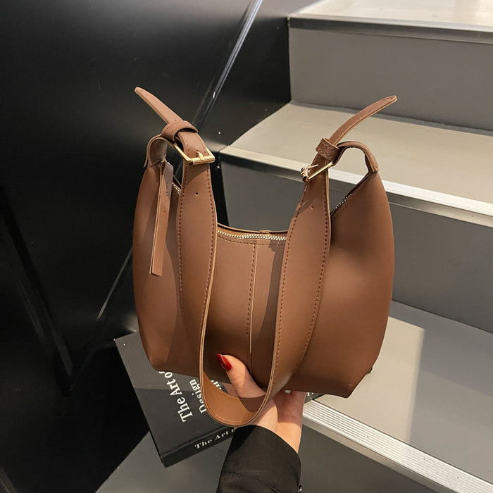A brown faux leather slouchy shoulder bag with a top zipper and adjustable shoulder straps, held by a hand with red nail polish against a modern black and gray staircase background.