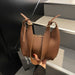 A brown faux leather slouchy shoulder bag with a top zipper and adjustable shoulder straps, held by a hand with red nail polish against a modern black and gray staircase background.