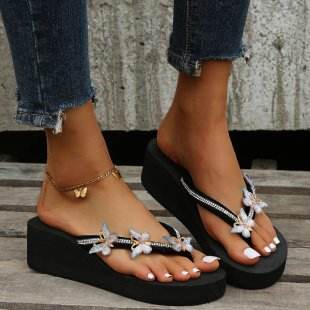 Black sandals with decorative straps worn by a person on a wooden floor.