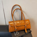A glossy tan leather handbag with double top handles and silver-tone hardware, displayed on a black book against a light gray textured wall.