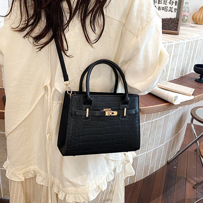A black crocodile-embossed handbag with gold-tone hardware, worn crossbody by a person in a cream-colored ruffled blouse in a cafe setting.