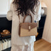 A woman viewed from behind wearing a structured handbag with a brown and beige color-blocked design, featuring a top handle, a shoulder strap, and a gold-tone turn-lock clasp, set against an indoor home background.