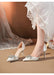 White high-heeled shoes with embellishments worn by a person in a white lace dress.
