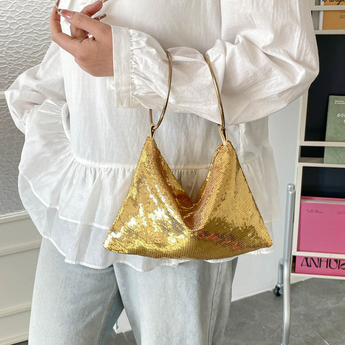 A person wearing a white ruffled blouse and light gray pants carries a small triangular gold sequin shoulder bag with a thin gold strap.