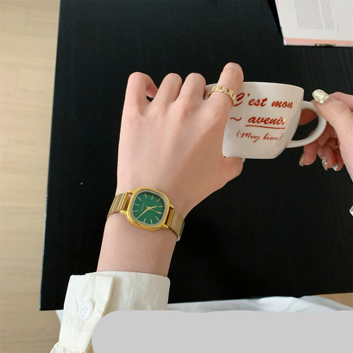 A gold-tone watch with a green dial and matching green hands is shown on a person's wrist, while they hold a white ceramic mug with red script text. The scene includes hands, a black tabletop, and part of a light wood floor, making it a lifestyle shot.