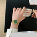 A gold-tone watch with a green dial and matching green hands is shown on a person's wrist, while they hold a white ceramic mug with red script text. The scene includes hands, a black tabletop, and part of a light wood floor, making it a lifestyle shot.