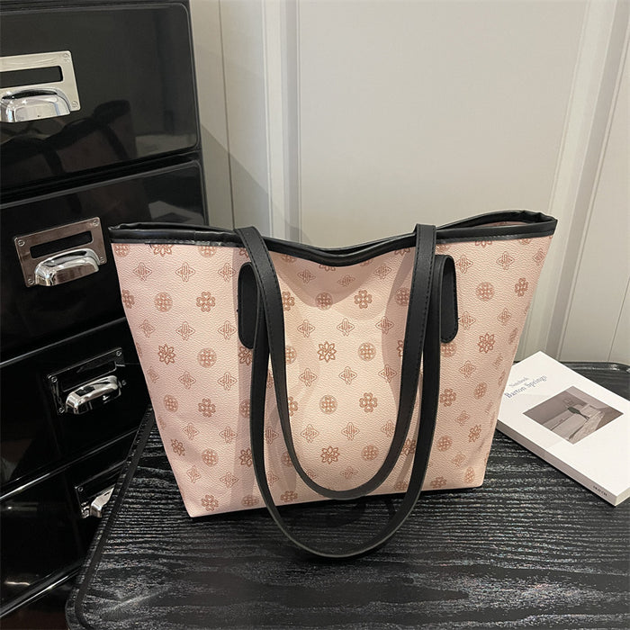 A light pink tote bag with a repeating brown geometric monogram pattern and black trim, resting on a dark wooden surface next to a black metal drawer and a white book.