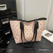 A light pink tote bag with a repeating brown geometric monogram pattern and black trim, resting on a dark wooden surface next to a black metal drawer and a white book.
