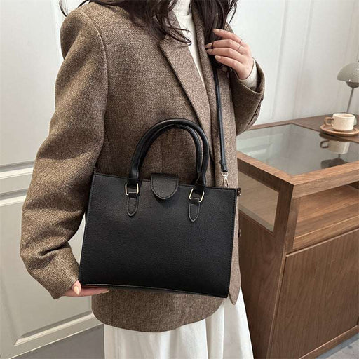 A woman wearing a brown blazer and white skirt carries a structured black leather handbag with double top handles and a shoulder strap in an indoor setting.