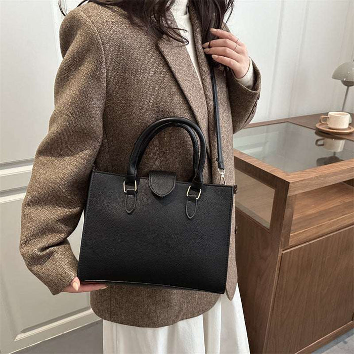 A woman wearing a brown blazer and white skirt carries a structured black leather handbag with double top handles and a shoulder strap in an indoor setting.