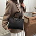 A woman wearing a brown blazer and white skirt carries a structured black leather handbag with double top handles and a shoulder strap in an indoor setting.