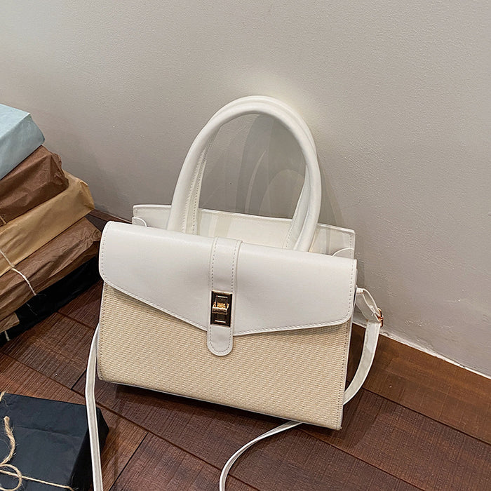 A structured handbag in off-white and cream, featuring a smooth faux-leather flap top with a gold-tone clasp, a woven lower body, dual top handles, and a detachable shoulder strap, resting on a wooden floor against a plain wall.