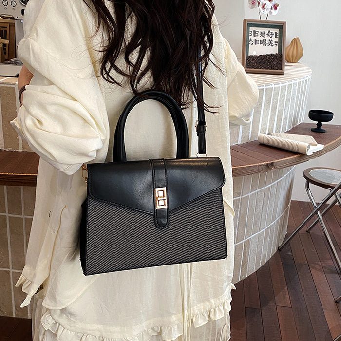A woman wearing a cream-colored blouse carries a structured black handbag with a top handle and a small gold accent on the front, set in a cafe environment.