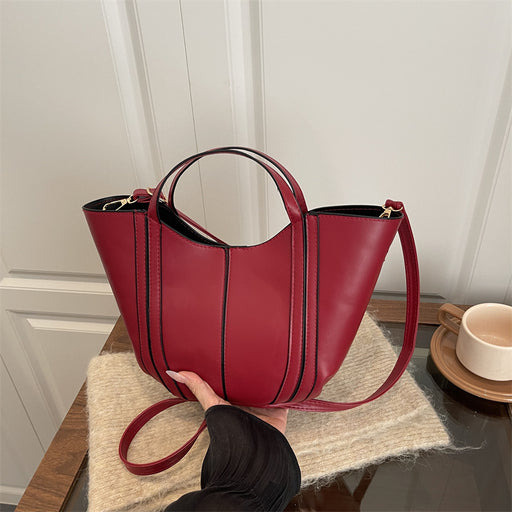 A red leather bucket tote bag with black vertical piping details, held by a hand over a beige textured mat on a wooden table next to a white cup.