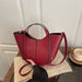 A red leather bucket tote bag with black vertical piping details, held by a hand over a beige textured mat on a wooden table next to a white cup.