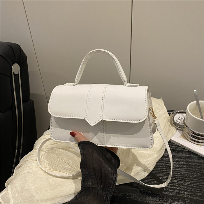 A white faux leather handbag with a top handle and shoulder strap, held by a hand wearing a black sleeve. The bag is resting on a white sheer fabric, with a black suitcase and a white cup in the background.