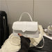 A white faux leather handbag with a top handle and shoulder strap, held by a hand wearing a black sleeve. The bag is resting on a white sheer fabric, with a black suitcase and a white cup in the background.